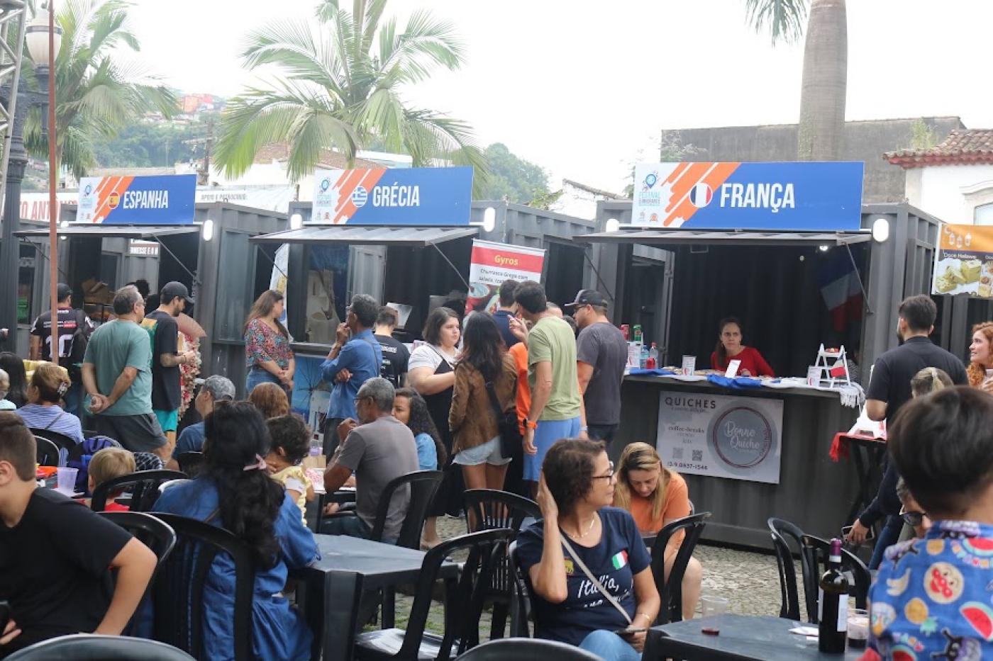 praça de alimentação com mesas ocupadas e estandes de alimentos onde se lê no alto de três deles: Espanha Grécia e França. #paratodosverem 