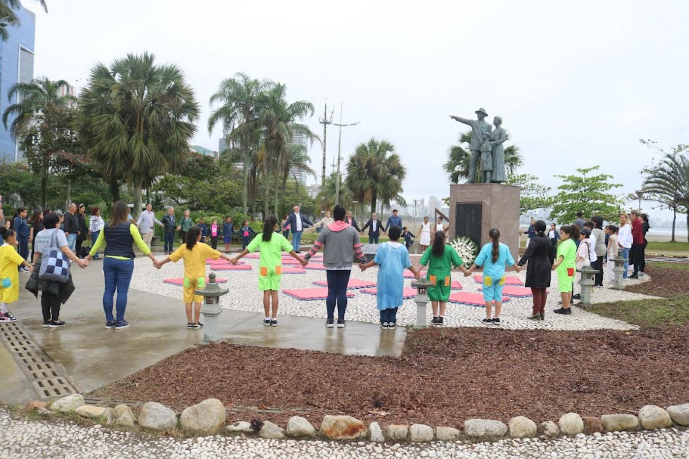 crianças e adultos dão as mãos em abraço simbólico em torno de monumento dedicado aos japoneses. #paratodosverm 