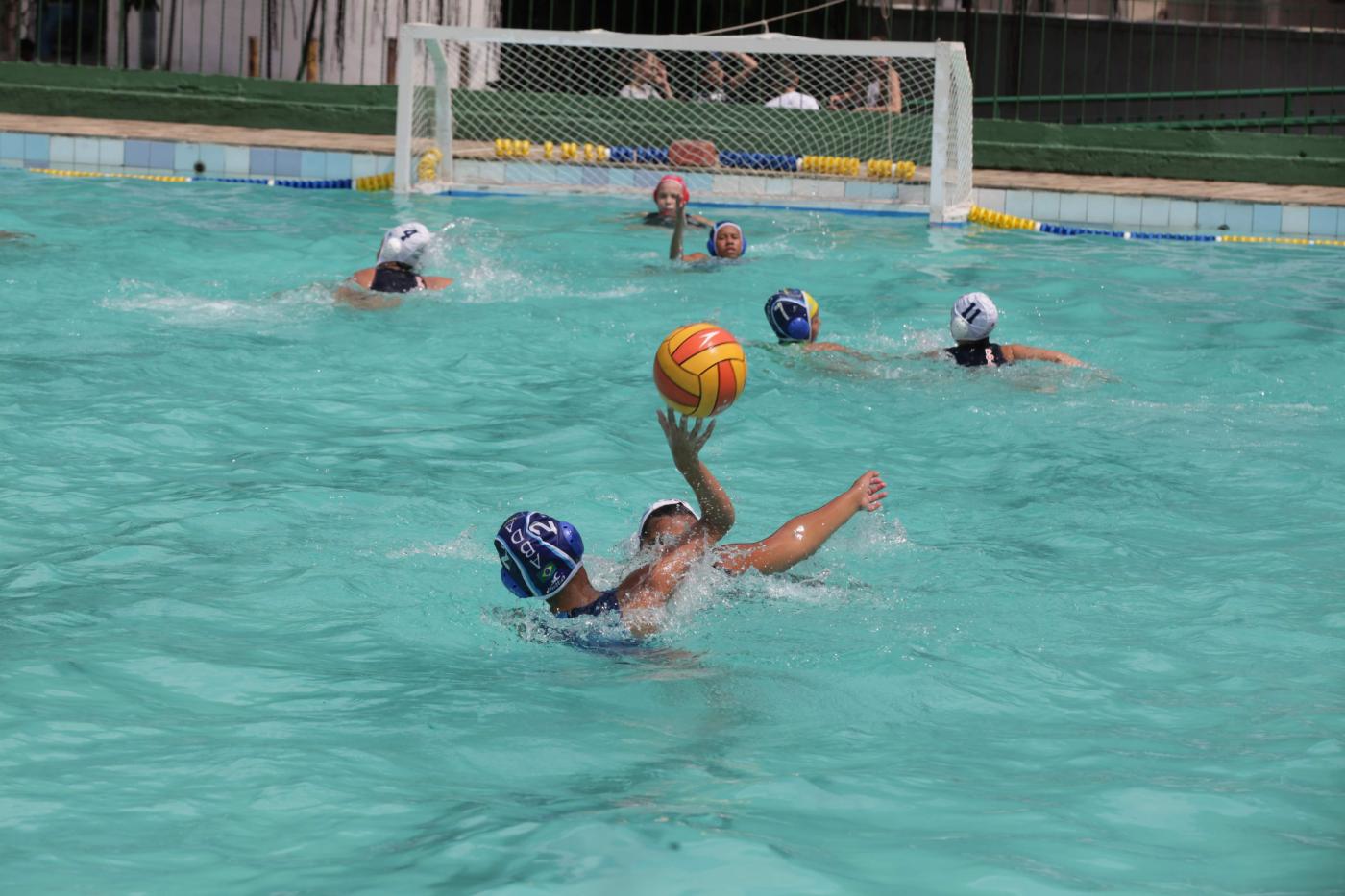 A piscina com atletas jogando polo aquático. Ao fundo está a trave. #Pracegover