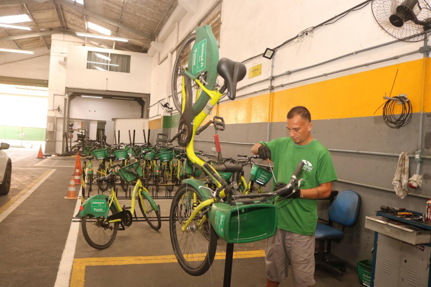 Homem conserta bicicleta. há bikes ao fundo do galpão. #paratodosverem