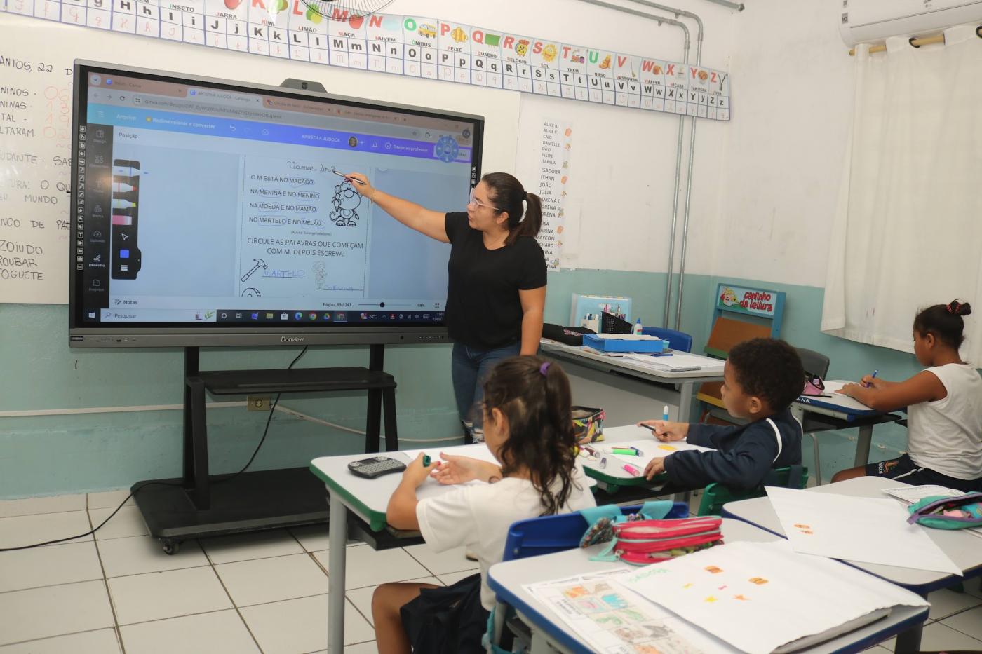 sala de aula com professora em pé falando aos alunos. #paratodosverem