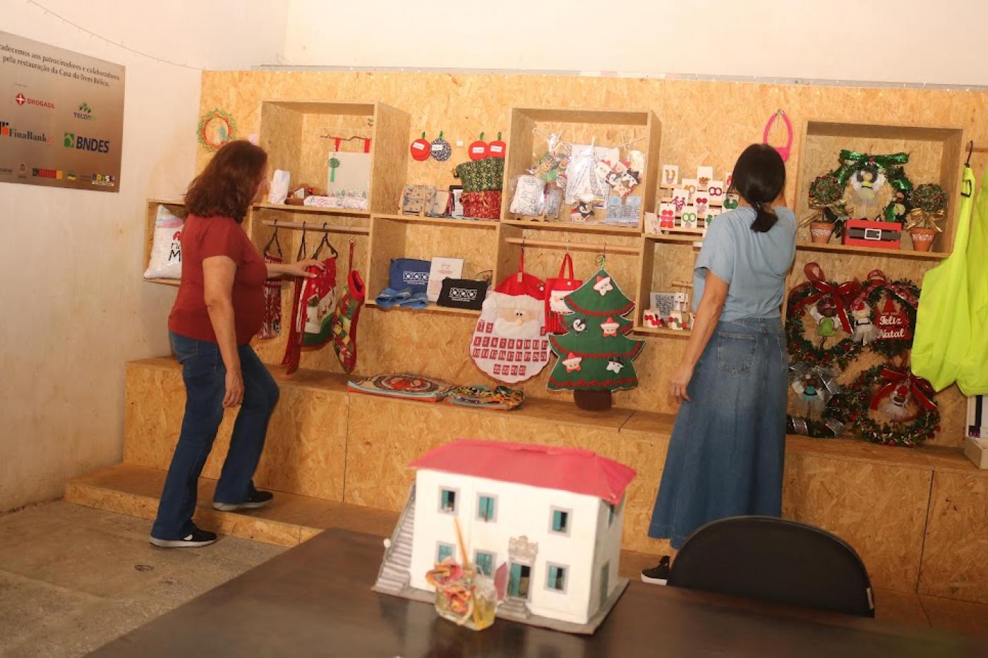 duas mulheres mexem em objetos artesanais expostos em prateleiras. Elas estão de costas para a foto. Em primeiro plano, uma miniatura da casa do trêm bélico sobre uma mesa. #paratodosverem 