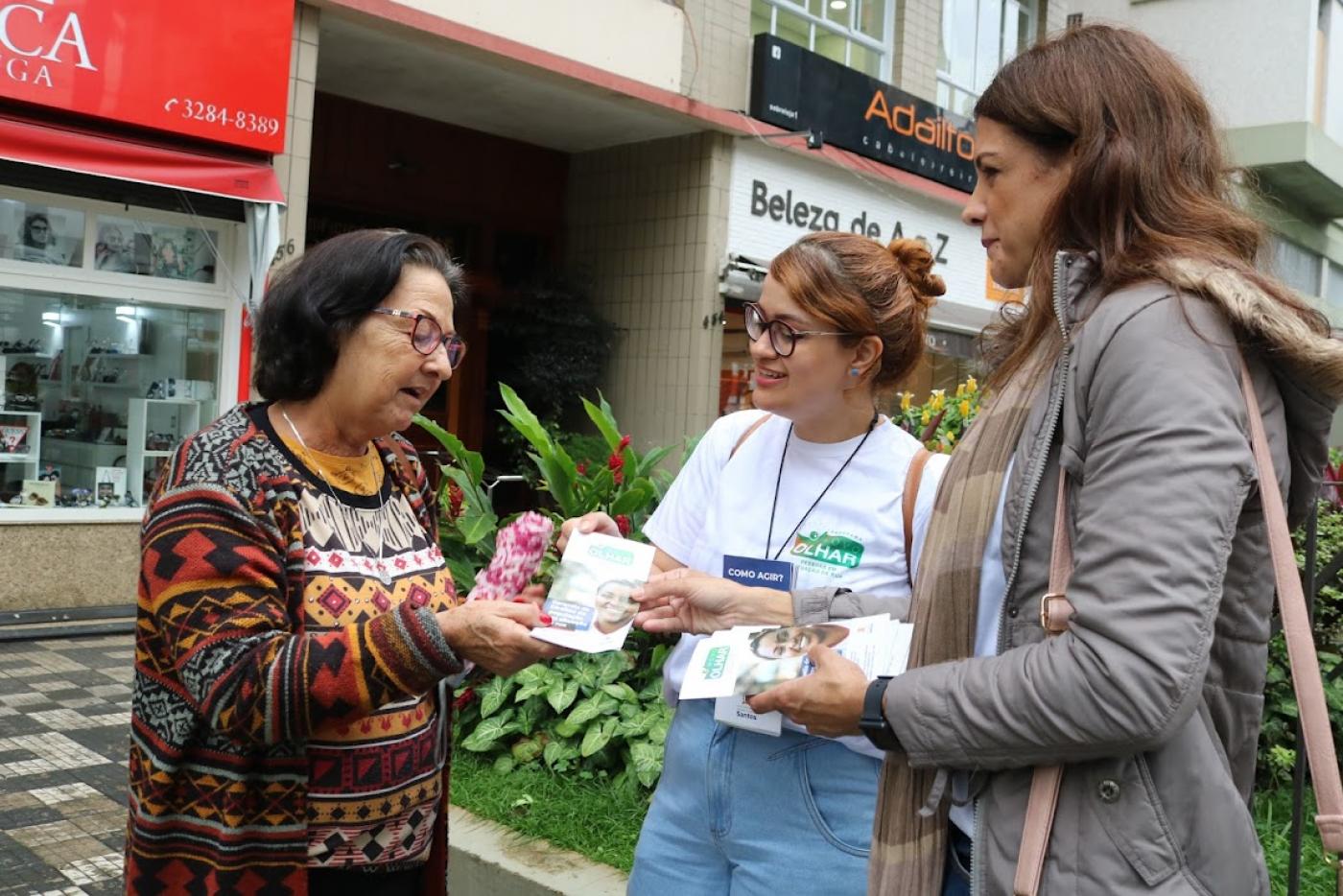 duas agentes entregam panfleto para mulher em calçada #paratodosverem