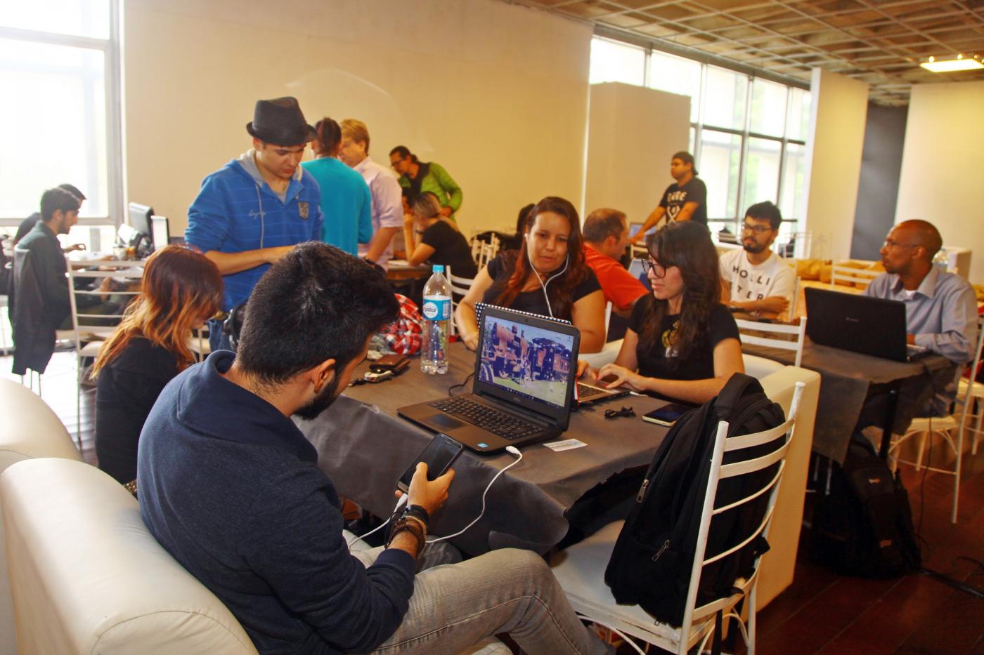 Jovens estão sentados em sofás, diante de mesas e na frente de seus notebooks. #Pracegover