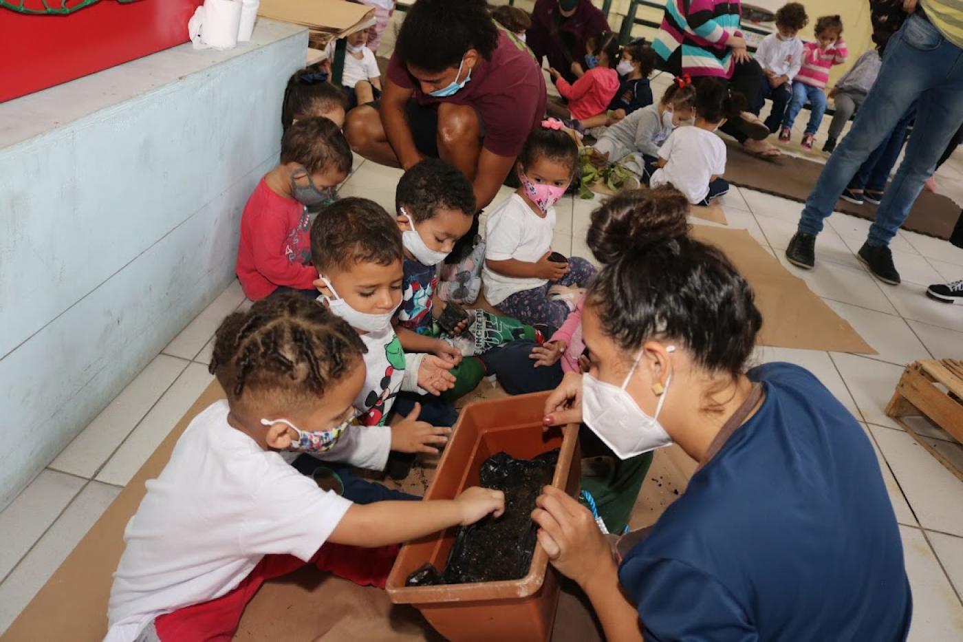 crianças com monitora colocam a mão em vaso com terra #paratodosverem