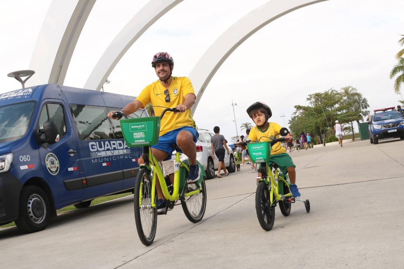 homem adulto e criança andam de bicicletas em parque. Ao lado deles está parada uma viatura da guarda municipal. #paratodosverem 