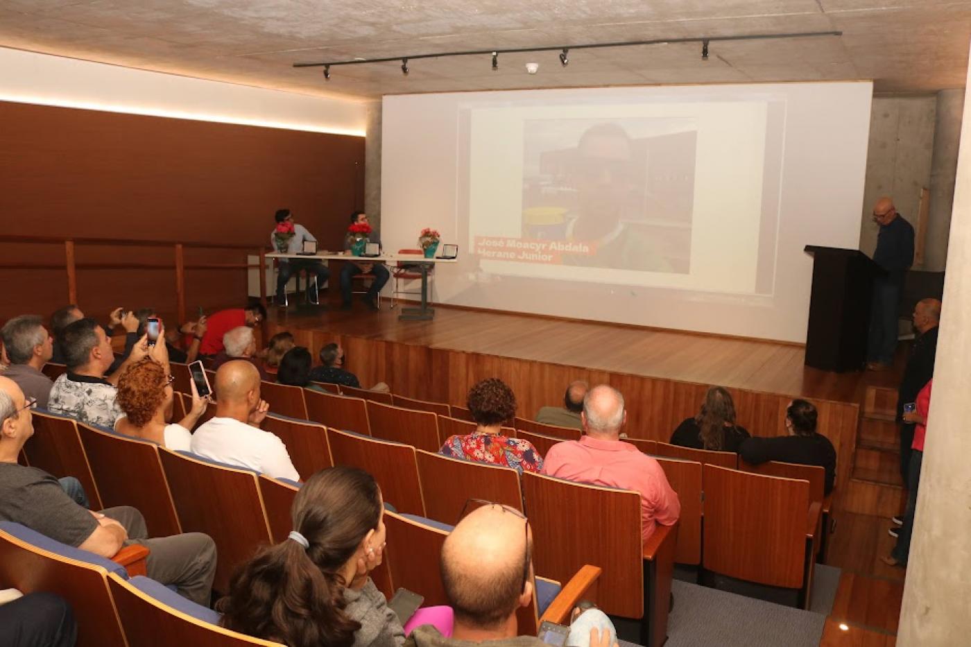 pessoas sentadas em auditório assistindo palestra e a imagens num telão ao fundo. #paratodosverem 