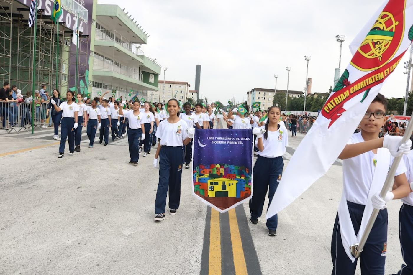 criança porta bandeira de santos em primeiro plano à direita. Ao fundo vem a banda escolar. #paratodosverem 