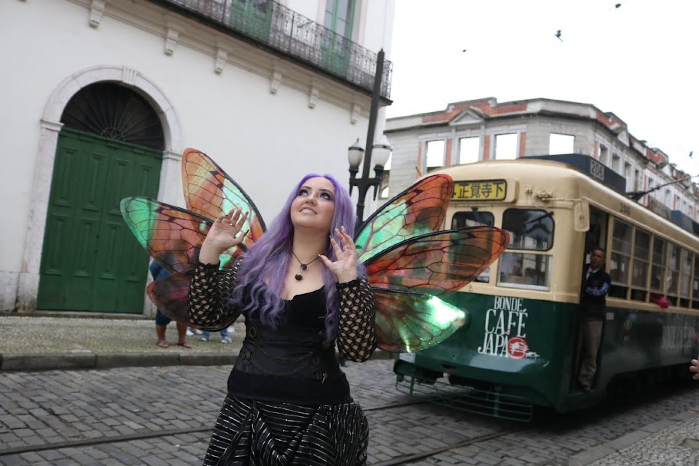 mulher com longo cabelo roxo usa asas de borboleta. O bonde está atrás dela. #paratodosverem 