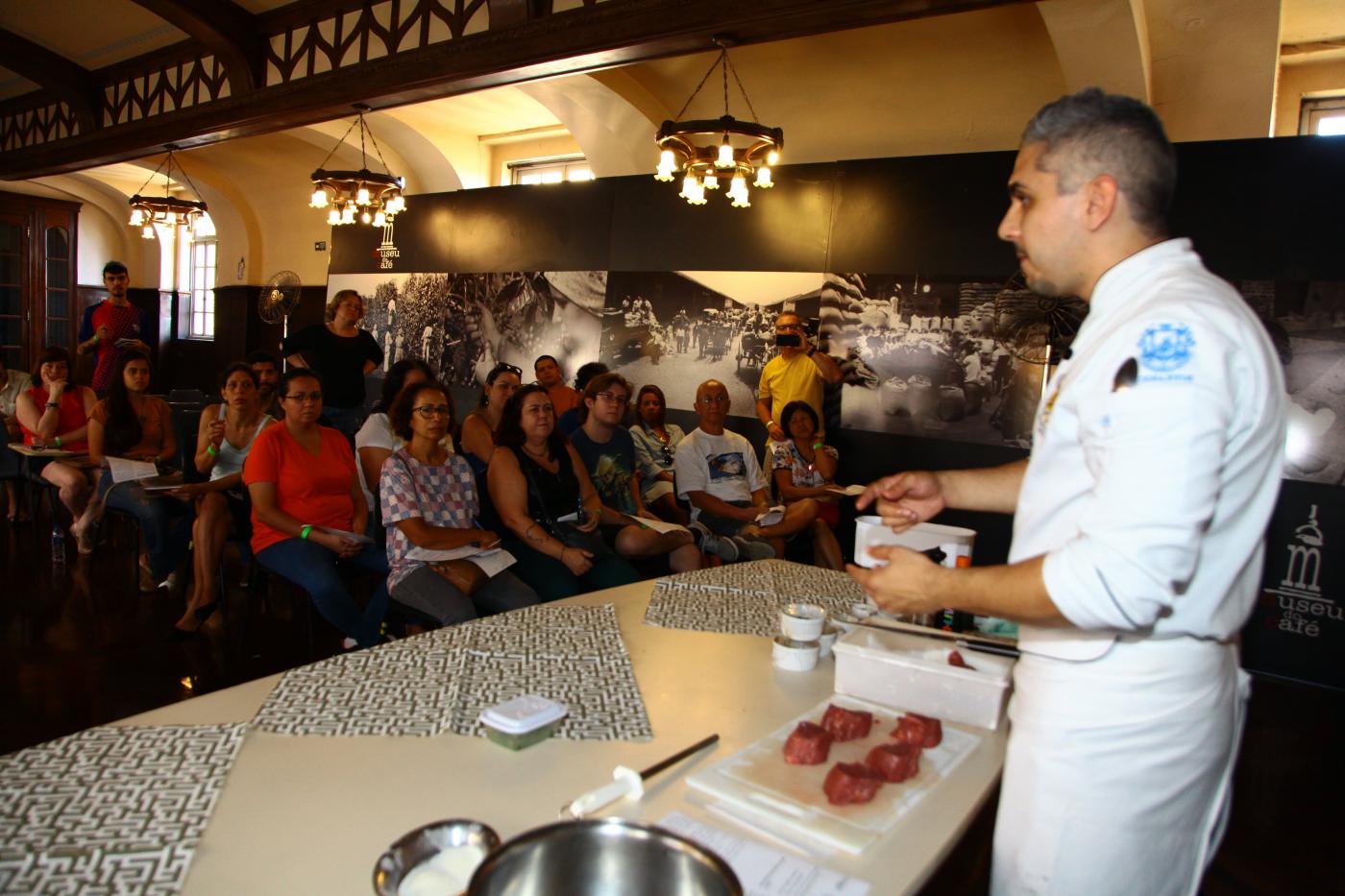 Aula de culinária dentro de sala de museu. O chefe está à frente de mesa, diante de uma tábua com alimentos. À frente está o público sentado assistindo a aula. #Pracegover