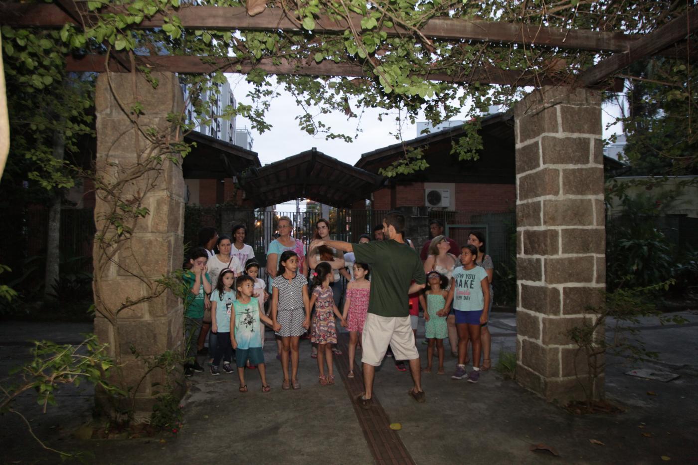 Crianças estão juntas na entrada de um parque. Monitor, de costas para a imagem, dá orientações ao grupo. Eles estão em meio à vegetação. #Pracegover