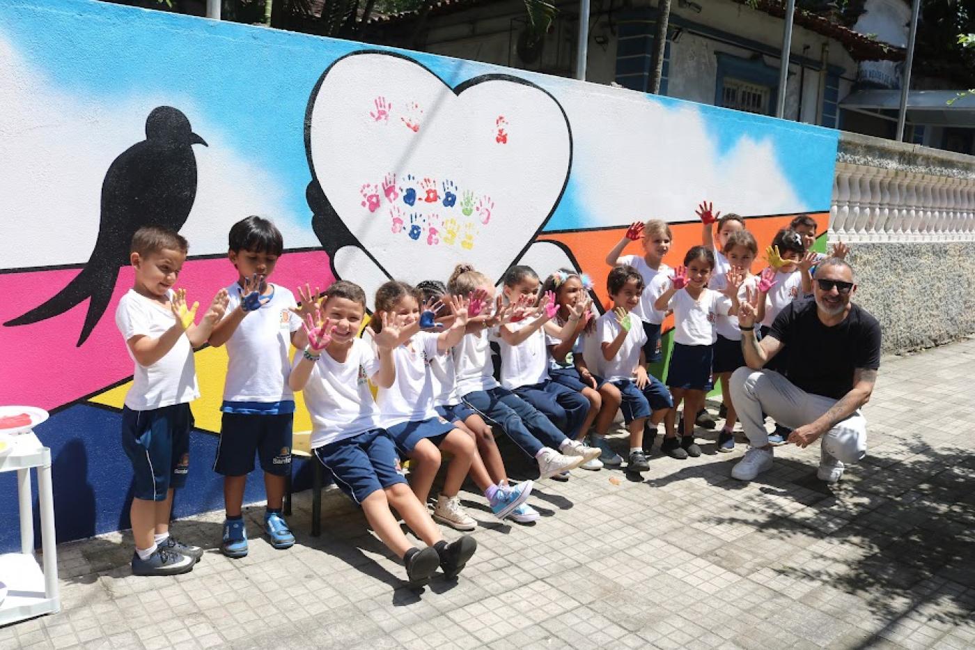 crianças estão sentadas em banco diante de arte pintada em muro. Elas esticam as mãos para a frente posando para a foto. #paratodosverem 