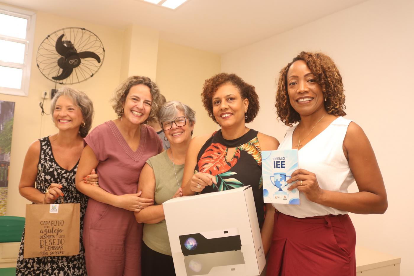 mulheres seguram pacote com projetor e azulejo decorado. #paratodosverem