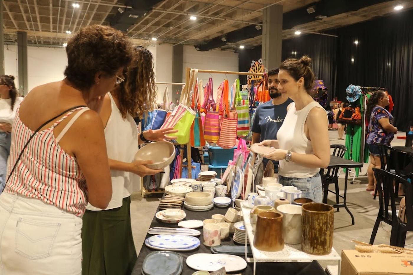 estande de venda de artesanato com duas consumidoras olhando os produtos e uma mulher, do outro lado da mesa, atendendo. #paratodosverem 