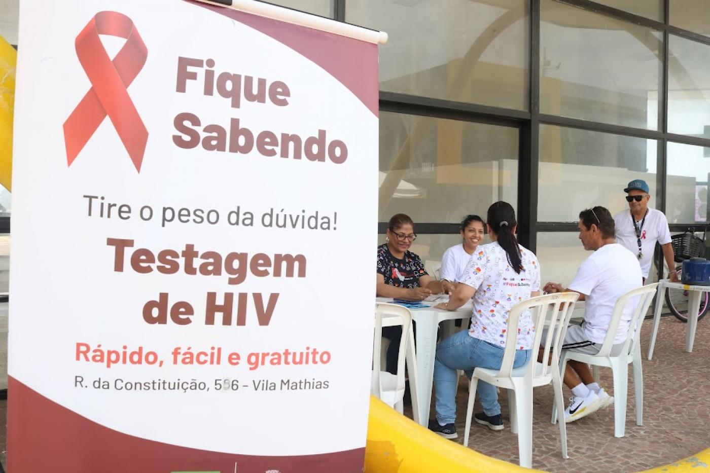 banner onde se lê Fique sabendo tire o peso da dúvida testagem de hiv. Ao fundo, pessoas fazem exames. #paratodosverem 
