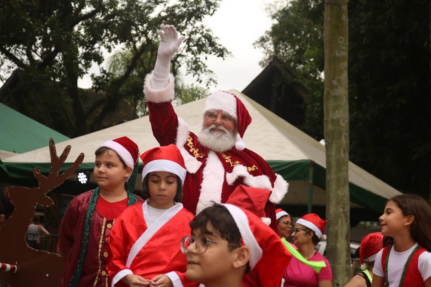 papai noel acena para o público. À crianças vestidas de vermelho e usando gorro à frente dele. #paratodosverem 