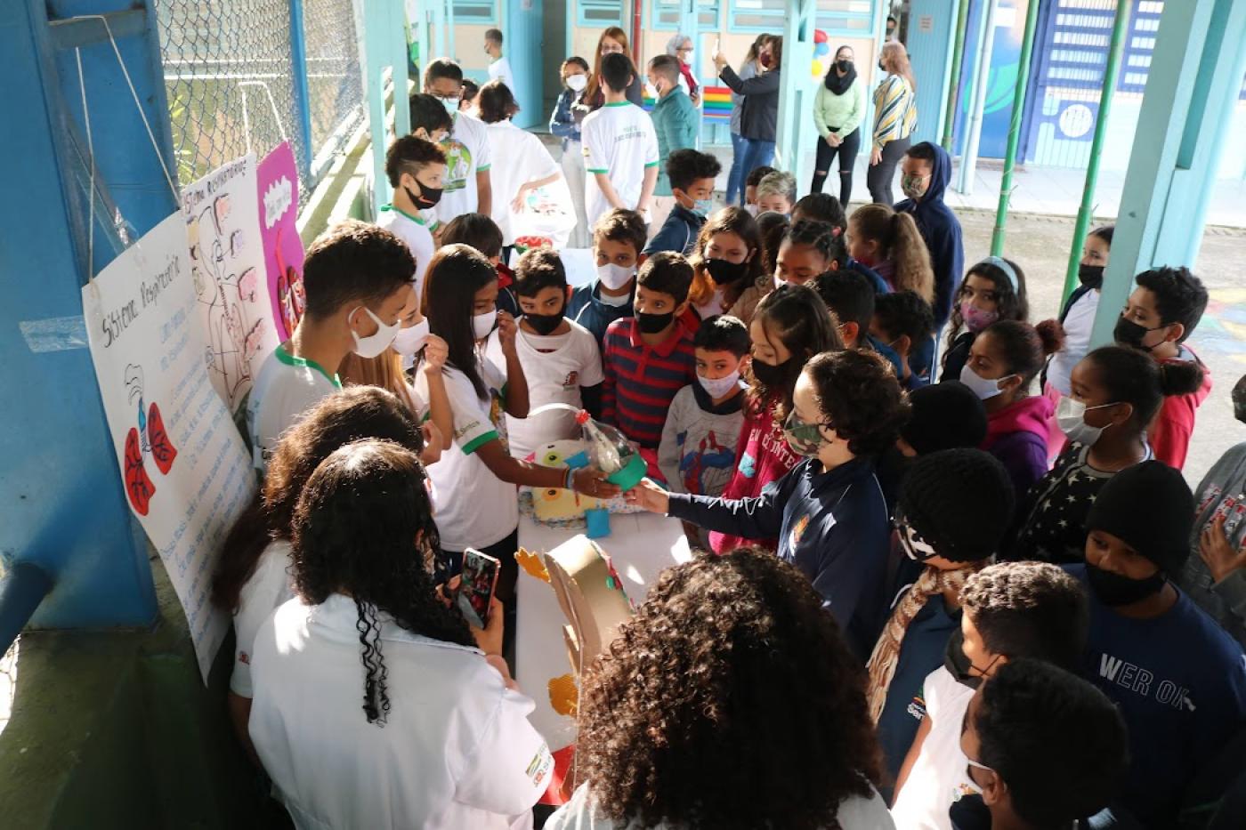 crianças cercam mesa com outros estudantes mostrando objetos #paratodosverem