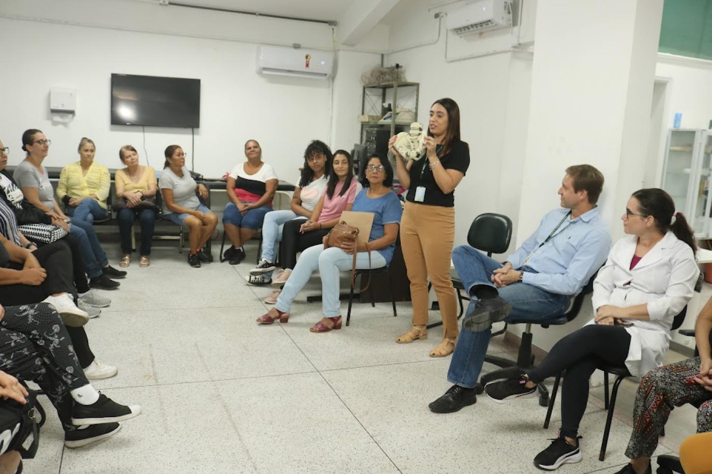 pessoas estão sentadas em círculo assistindo palestra. uma mulher fala em pé ao grupo. #paratodosverem