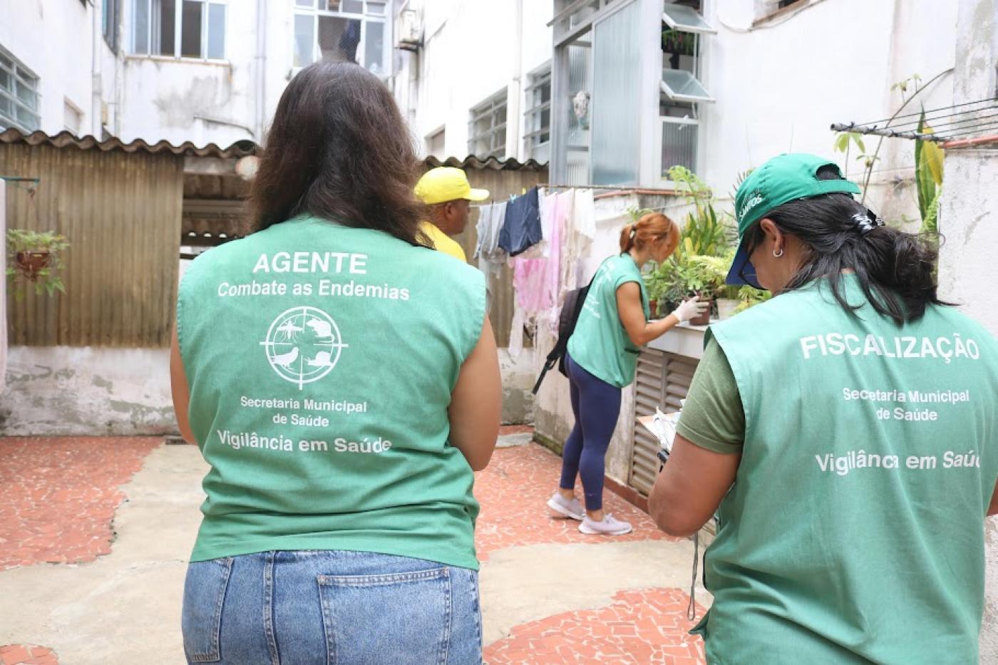 duas agentes da dengue uniformizadas, de costas para a foto. Ao fundo uma agente vistoria vaso em área aberta de uma casa. #paratodosverem 
