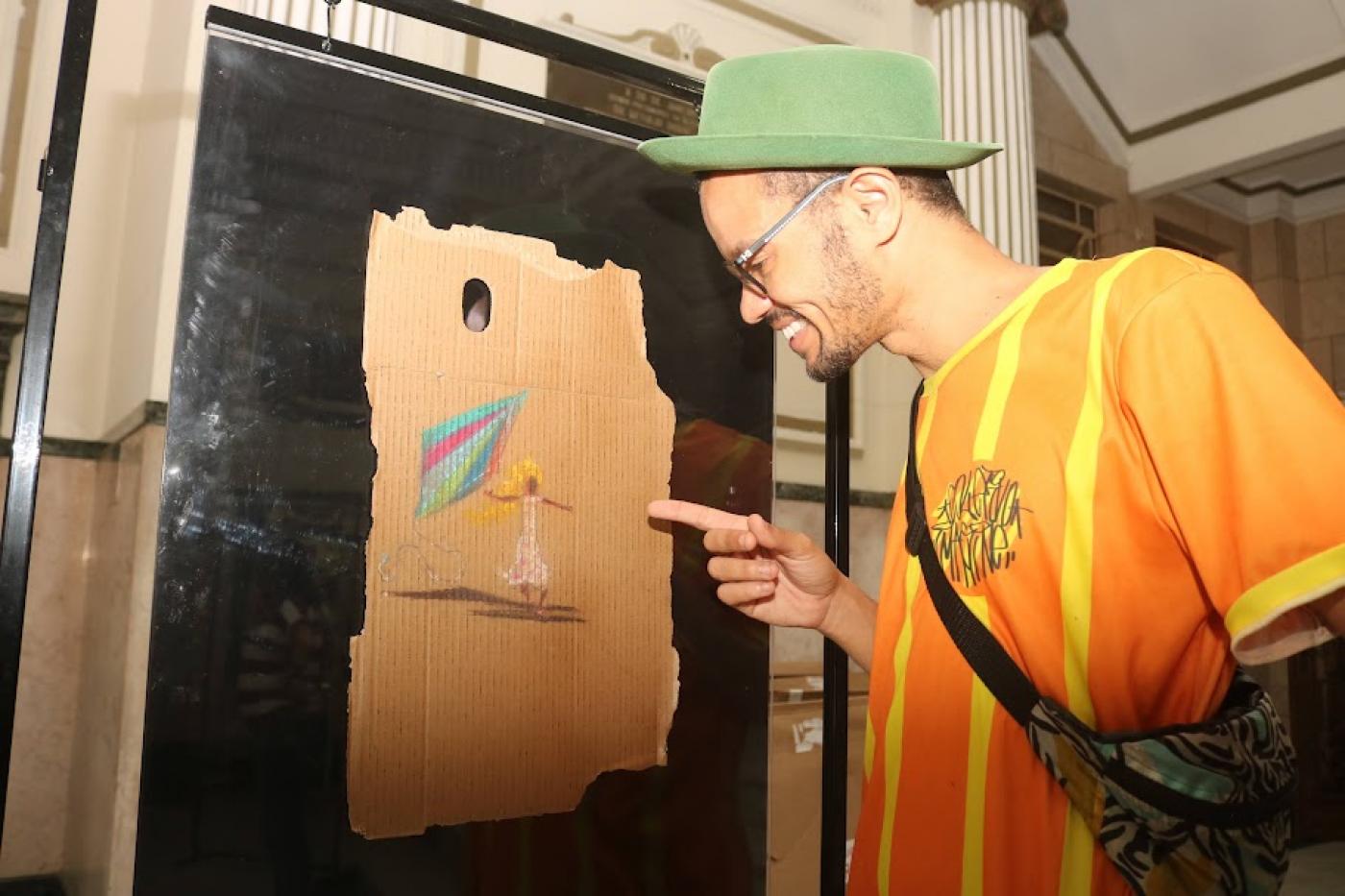 artista aponta para uma de suas obras afixada em display. #paratodosverem 