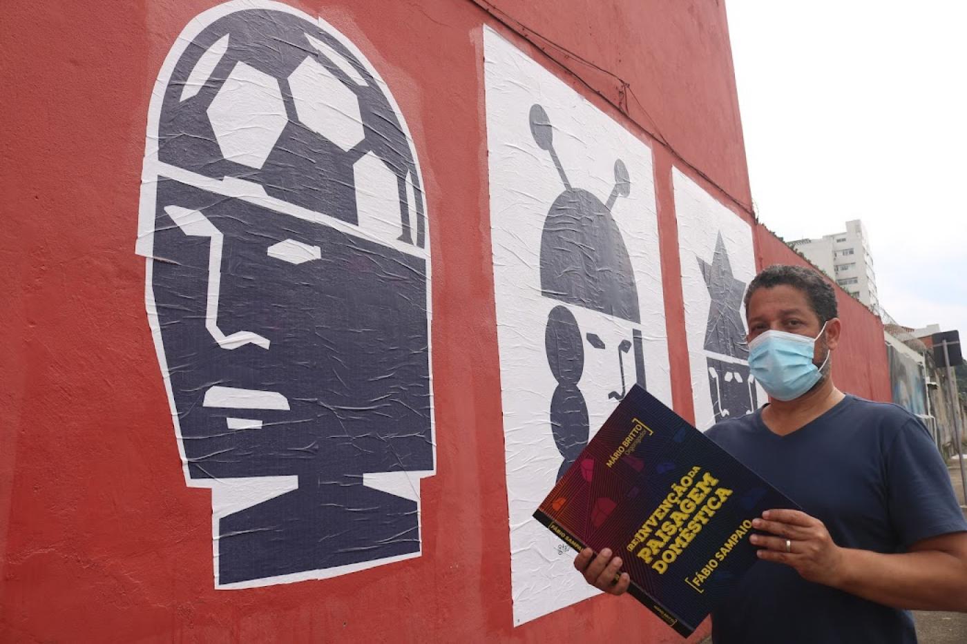 Artista segura livro em frente de parede com colagens #paratodosverem