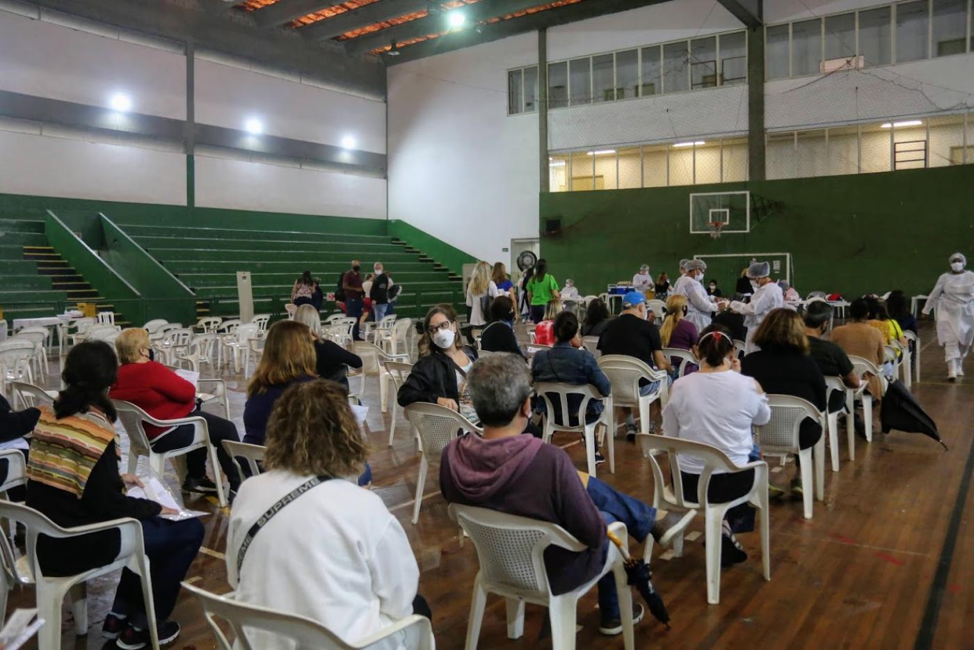 Pessoas estão sentadas em cadeiras afastadas em ginásio esportivo. #paratodosverem