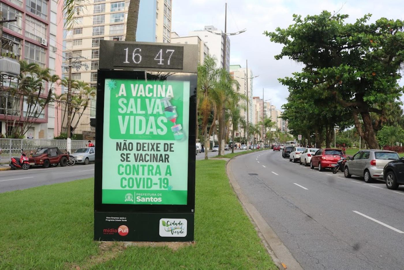 toten com relógio informando a hora no topo. Abaixo, orientação sobre vacina. Toten está em canteiro central na avenida da oral. Os prédios estão do lado esquerdo. A pista à direita está sem movimento, somente com veículos estacionados. #paratodosverem