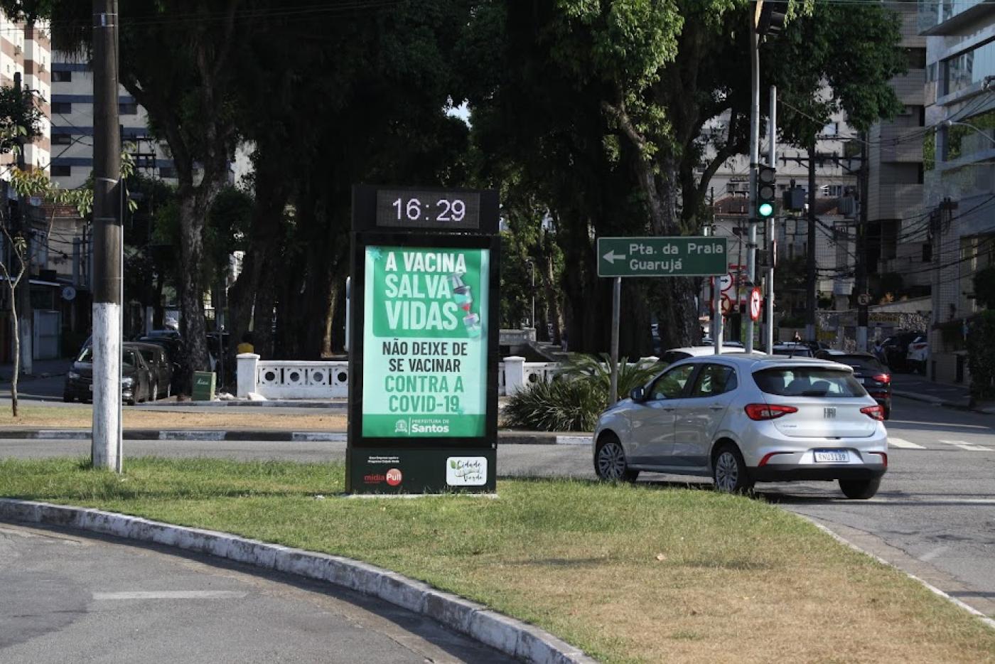 Totem em canteiro central com horário marcando 16h29. O restante do totem tem informação sobre a importância da vacina. Há um carro fazendo curva ao lado. #paratodosverem
