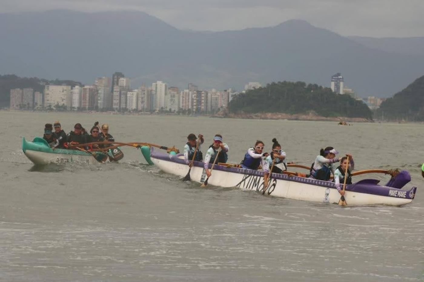 duas canoas dirigidas por mulheres estão no mar. A que está atrás tenta alcançar a que está à frente. #paratodosverem