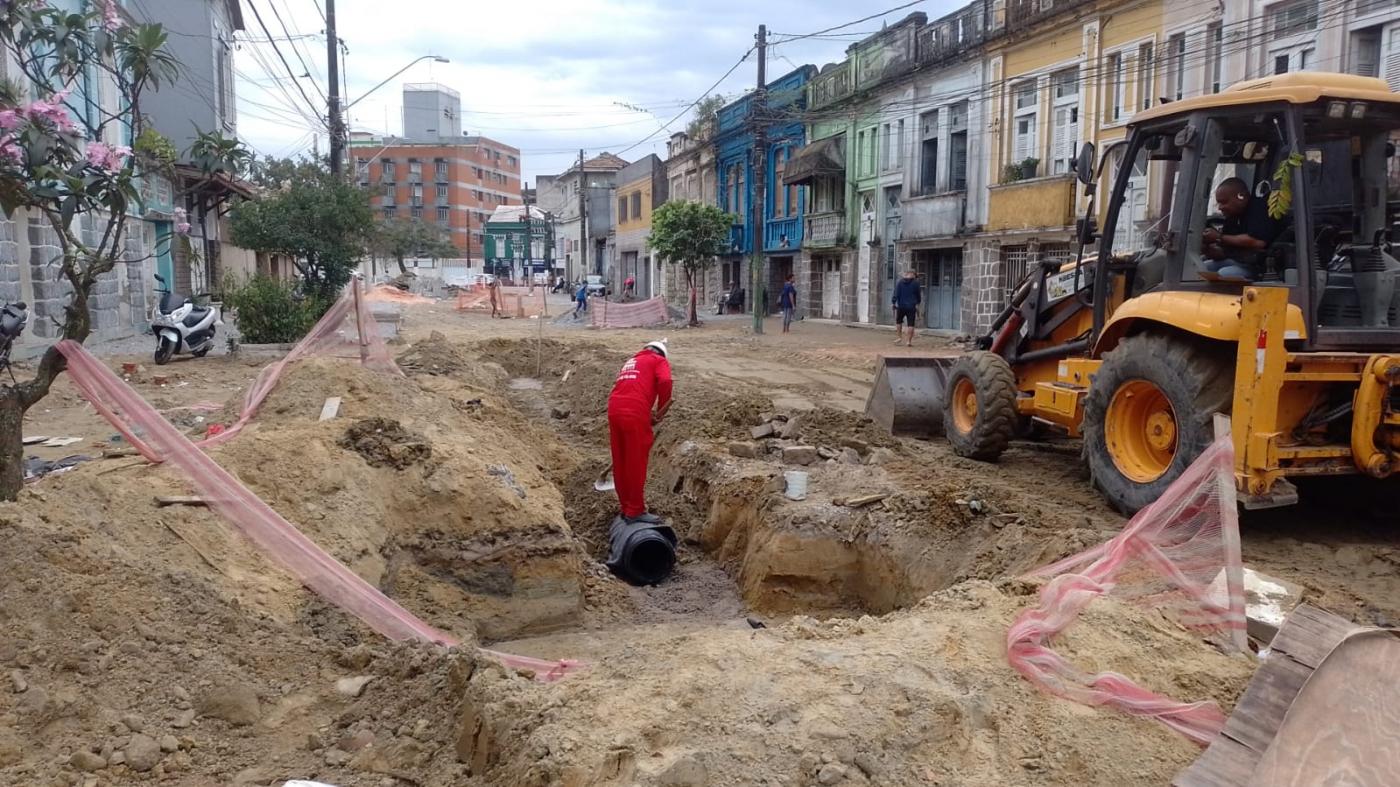 homem está trabalhando em escavação no leito da rua. Ao lado direito dele há um trator. #paratodosverem