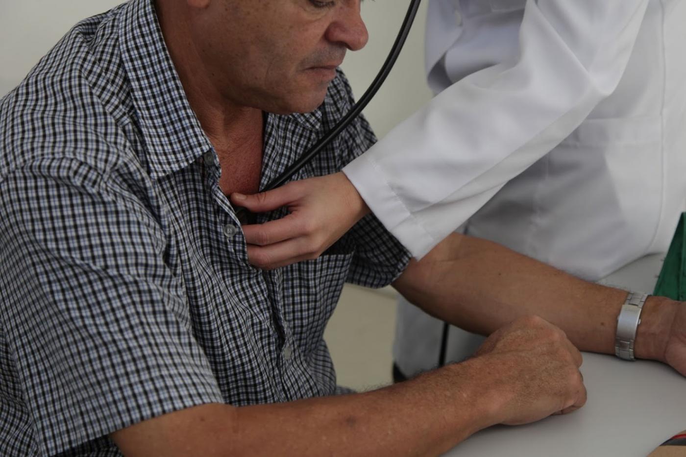 Foto em cluse, com homem sendo auscultado. Mão que segura o 