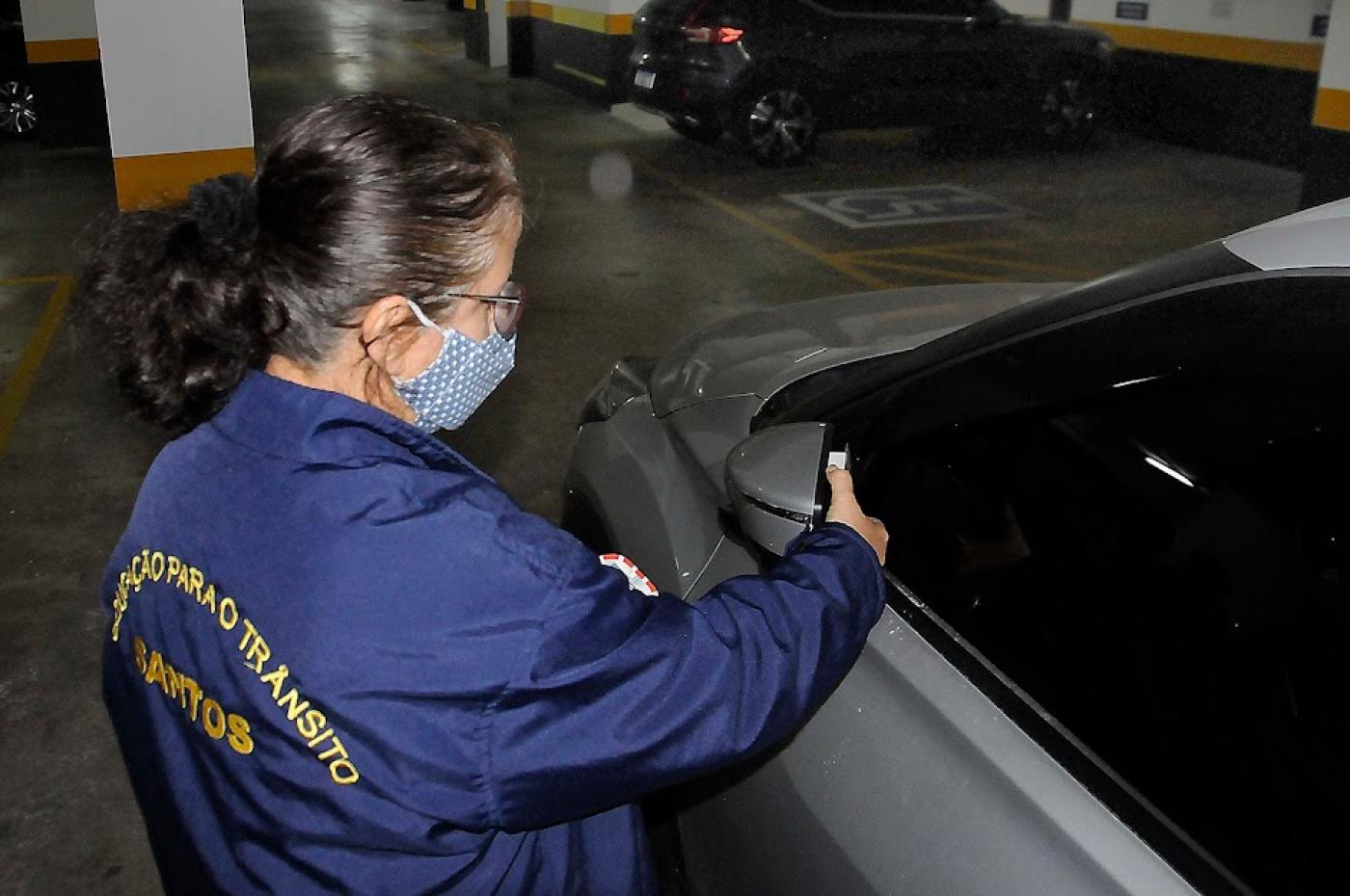 Agente afixa informativo em retrovisor de carro em estacionamento. #pracegover