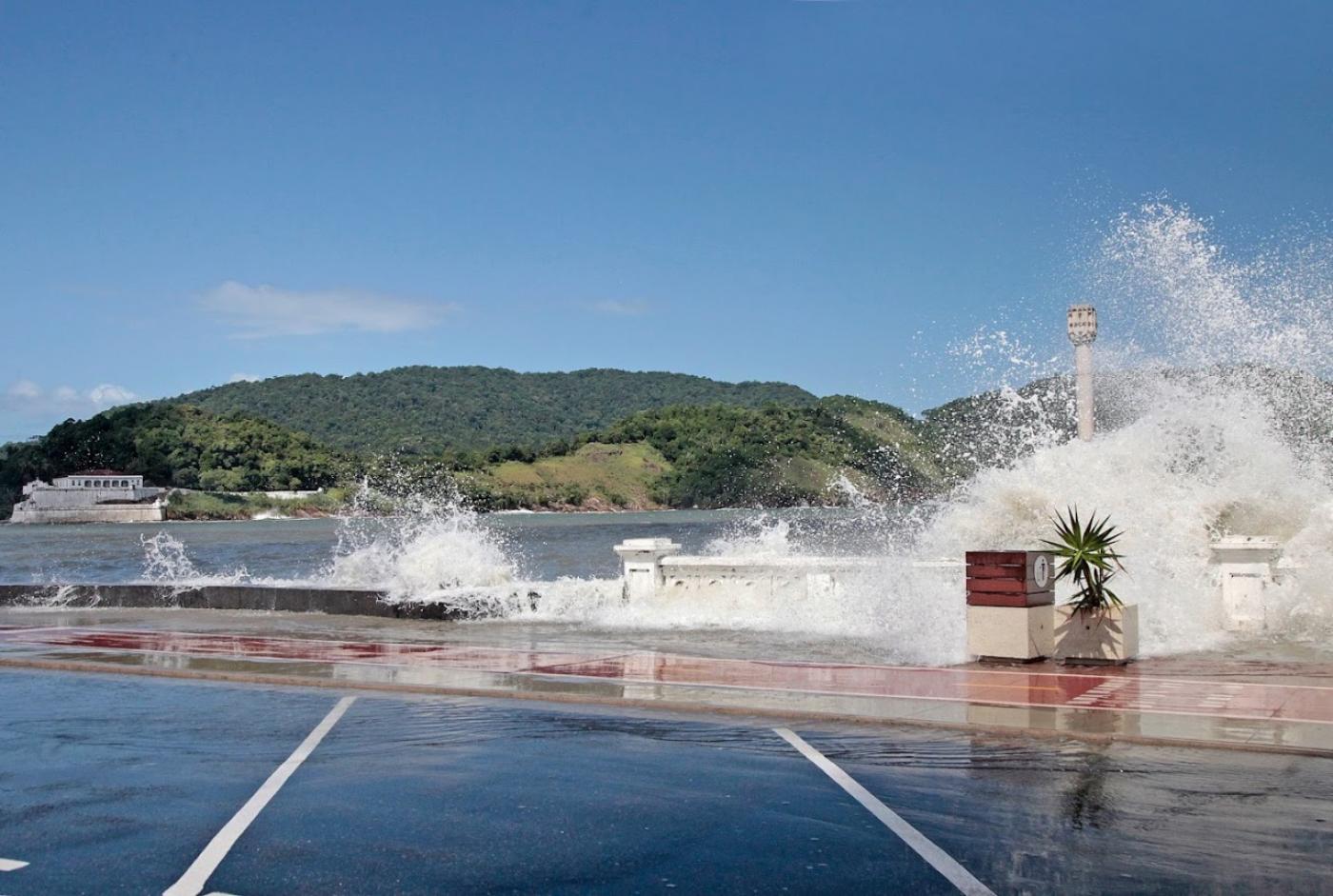 Em ressaca, ondas do mar batem na mureta da ponta da praia. #paratodosverem