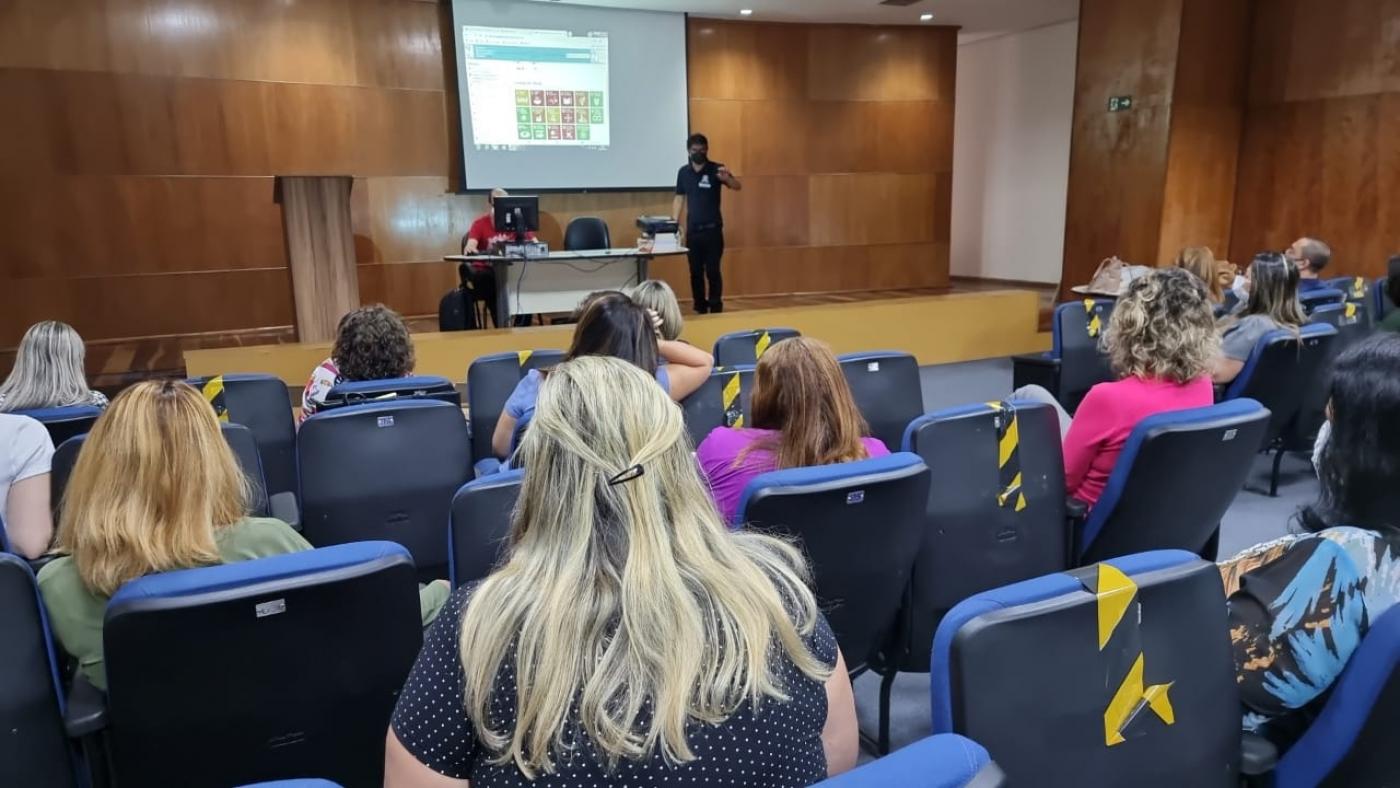 Pessoas sentadas em auditório assistindo palestrante #paratodosverem