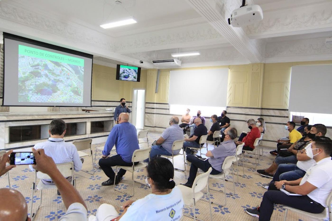 pessoas estão sentadas em cadeiras distribuídas em salão. Todas assistem a uma projeção num telão e estão atentas a um homem que fala à frente de todos.#paratodosverem