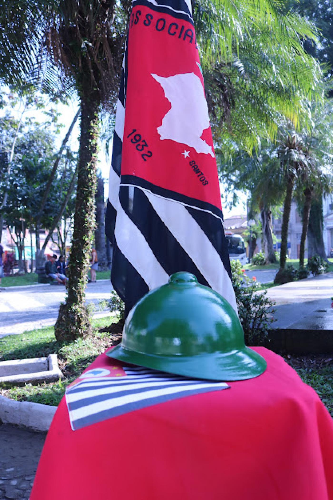 Modelo de capacete verde, dos antigos combatentes, sobre bandeira de São Paulo em mesa com toalha vermelha. #pratodosverem