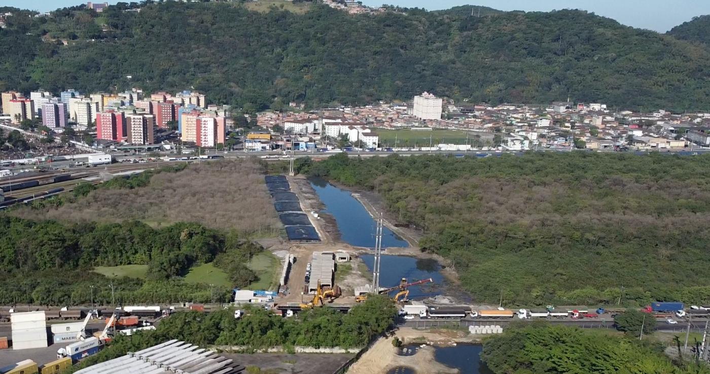 imagem área aparece rio com máquinário de obras ao lado, vasta vegetação ao redor e prédios e morro ao fundo #paratodosverem