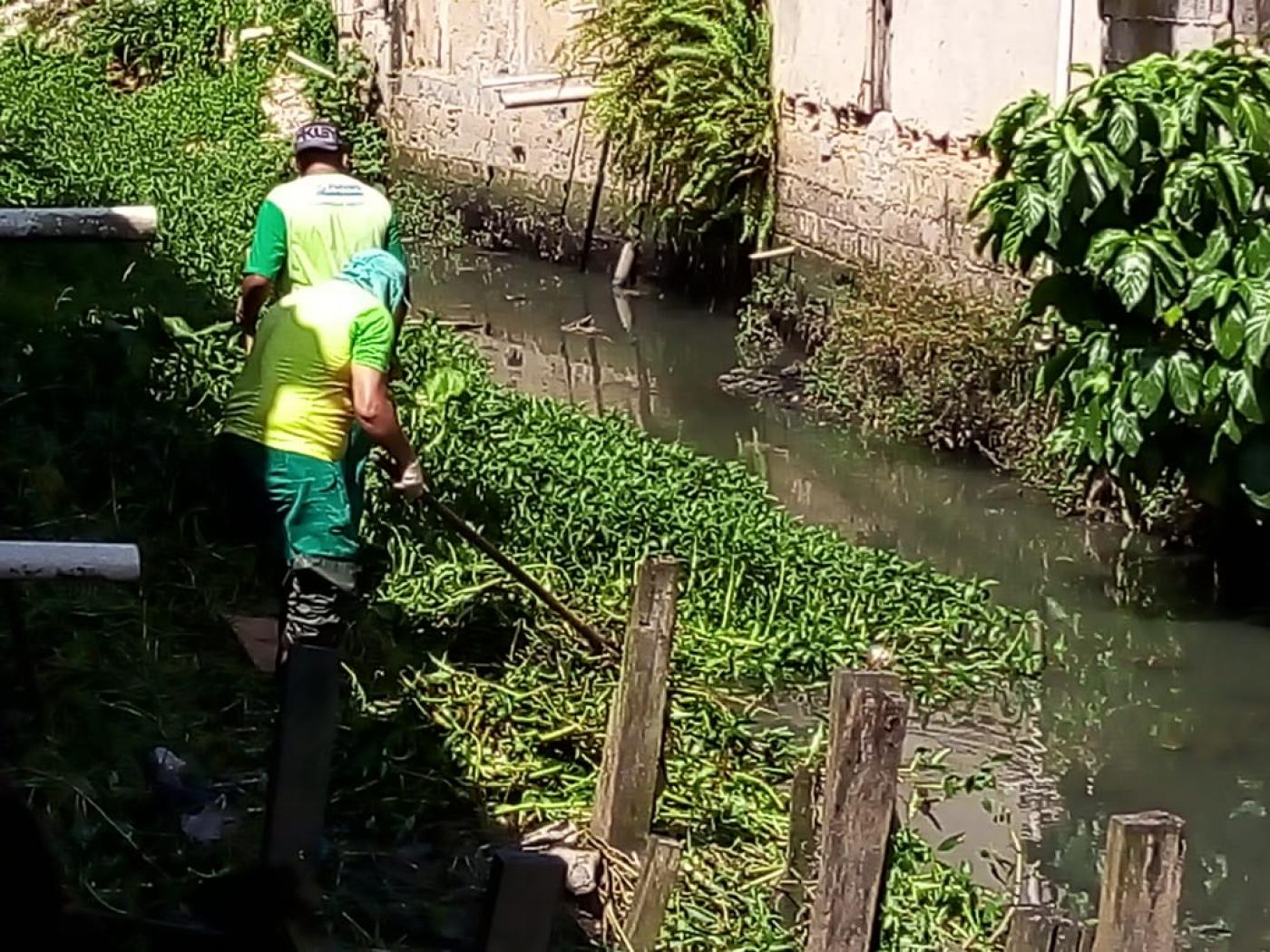 Dois trabalhadores removem resíduos em margem de rio. #Paratodosverem