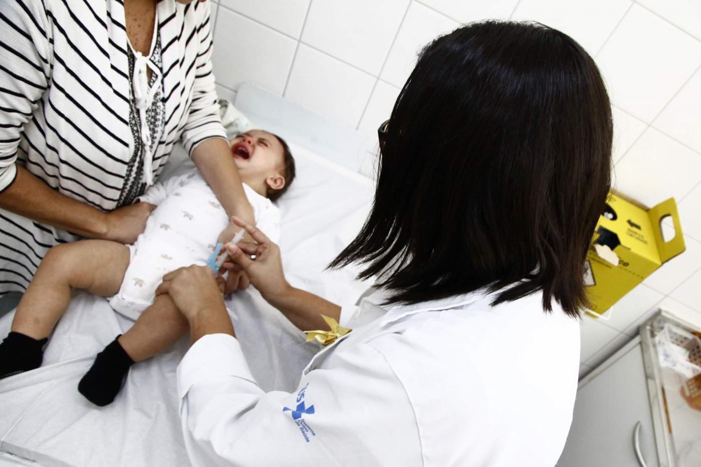 Criança está deitada em maca sendo vacinada. As mãos de uma mulher seguram o bebê. #Pracegover