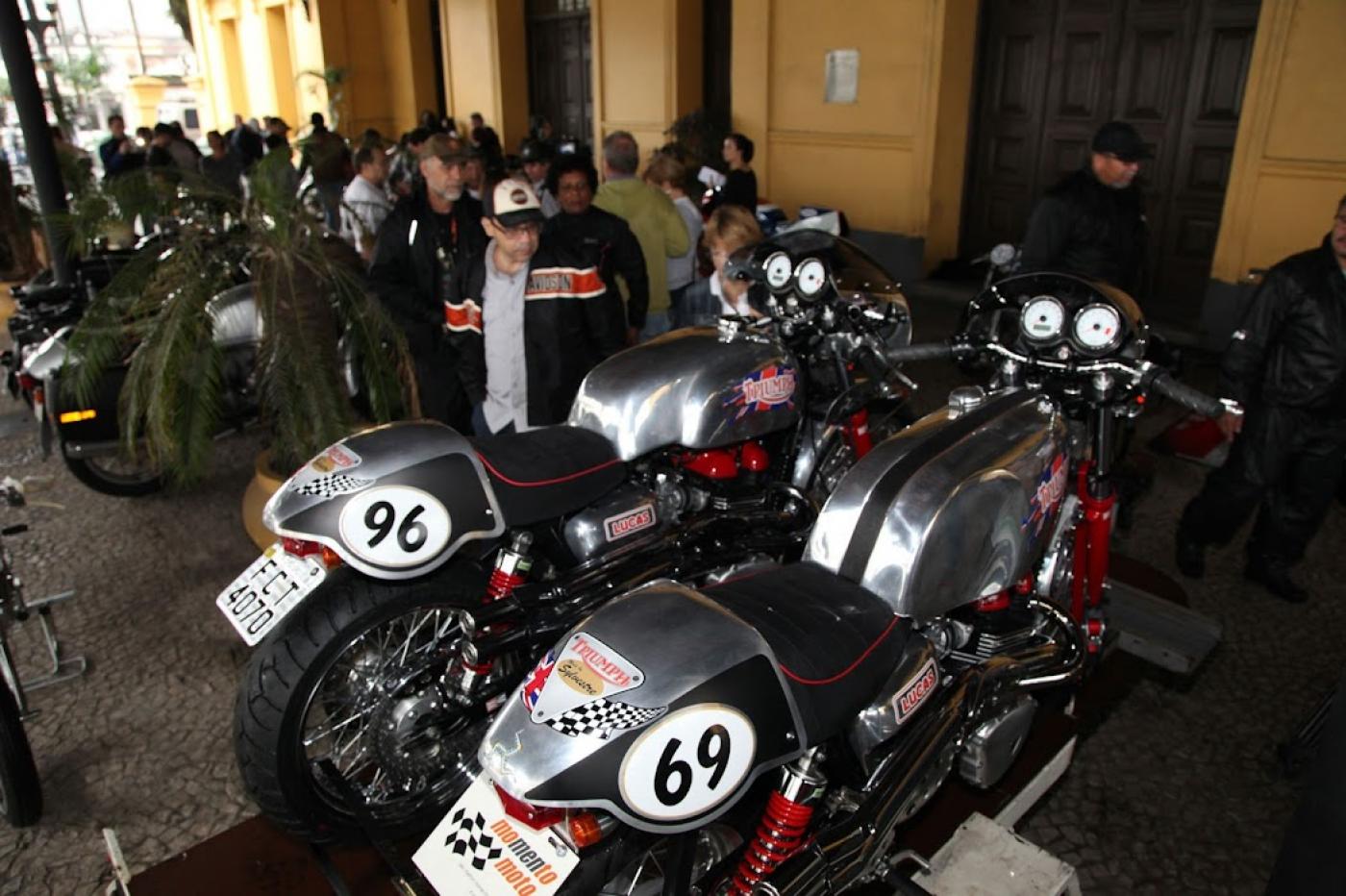 Duas motos antigas na frente de estação e várias pessoas ao fundo #paratodosverem