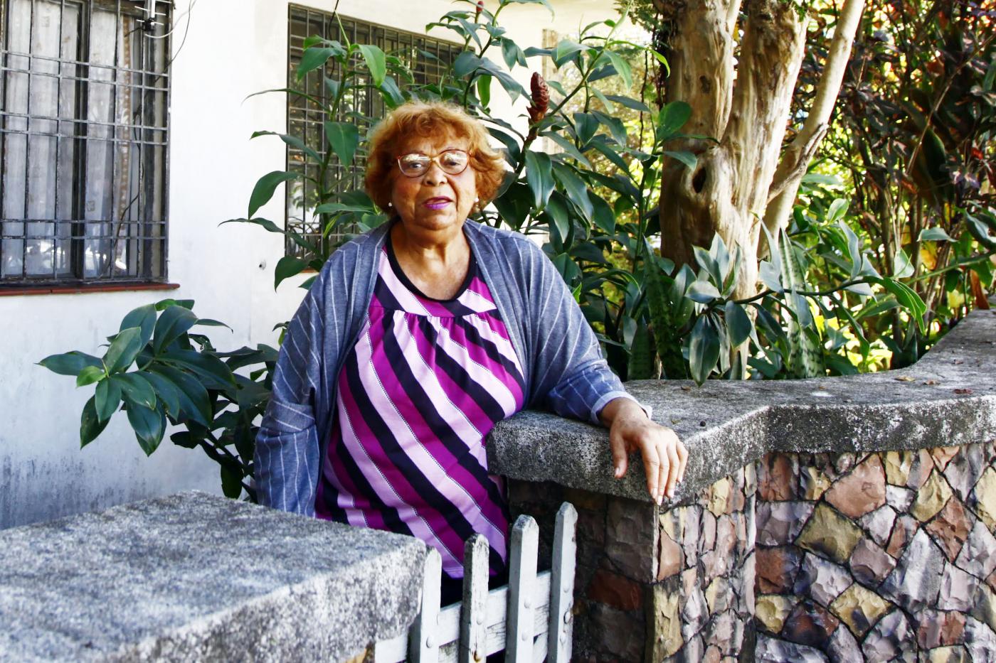 Leonita posa para foto apoiada em mureta da frente da casa, feita de pedras, com cerca de um metro de altura