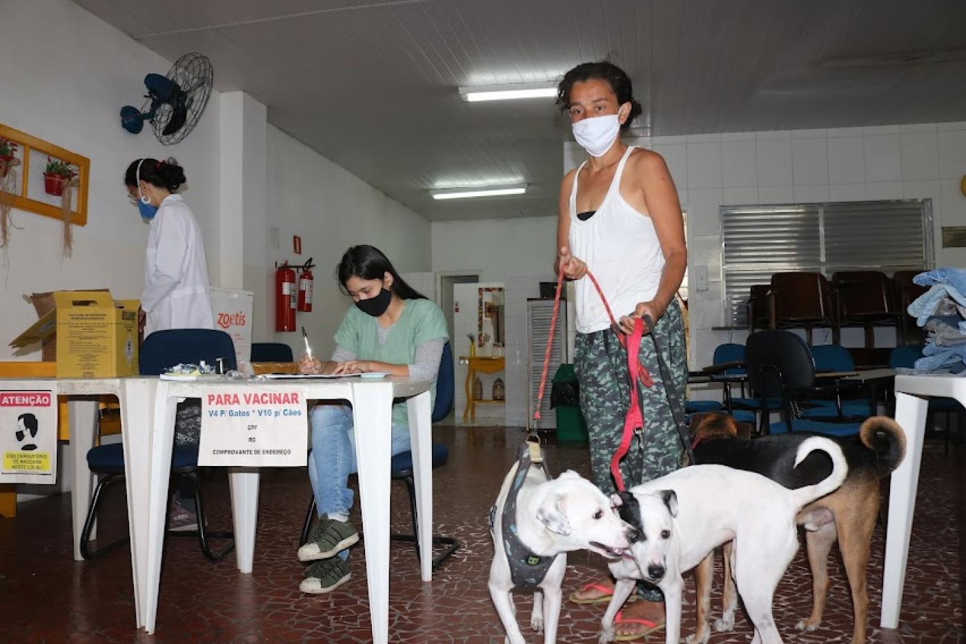 Mulher segura dois cachorros na coleira, enquanto mulher está sentada fazendo anotações. #paratodosverem