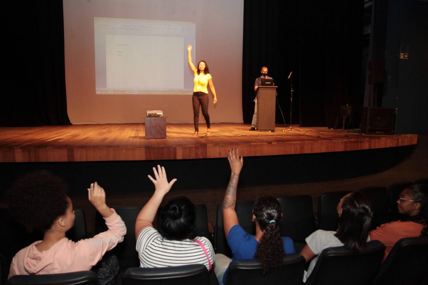 em um palco mulher com mão levantada, na platéia , de costas mulheres sentadas e com mãos levantadas