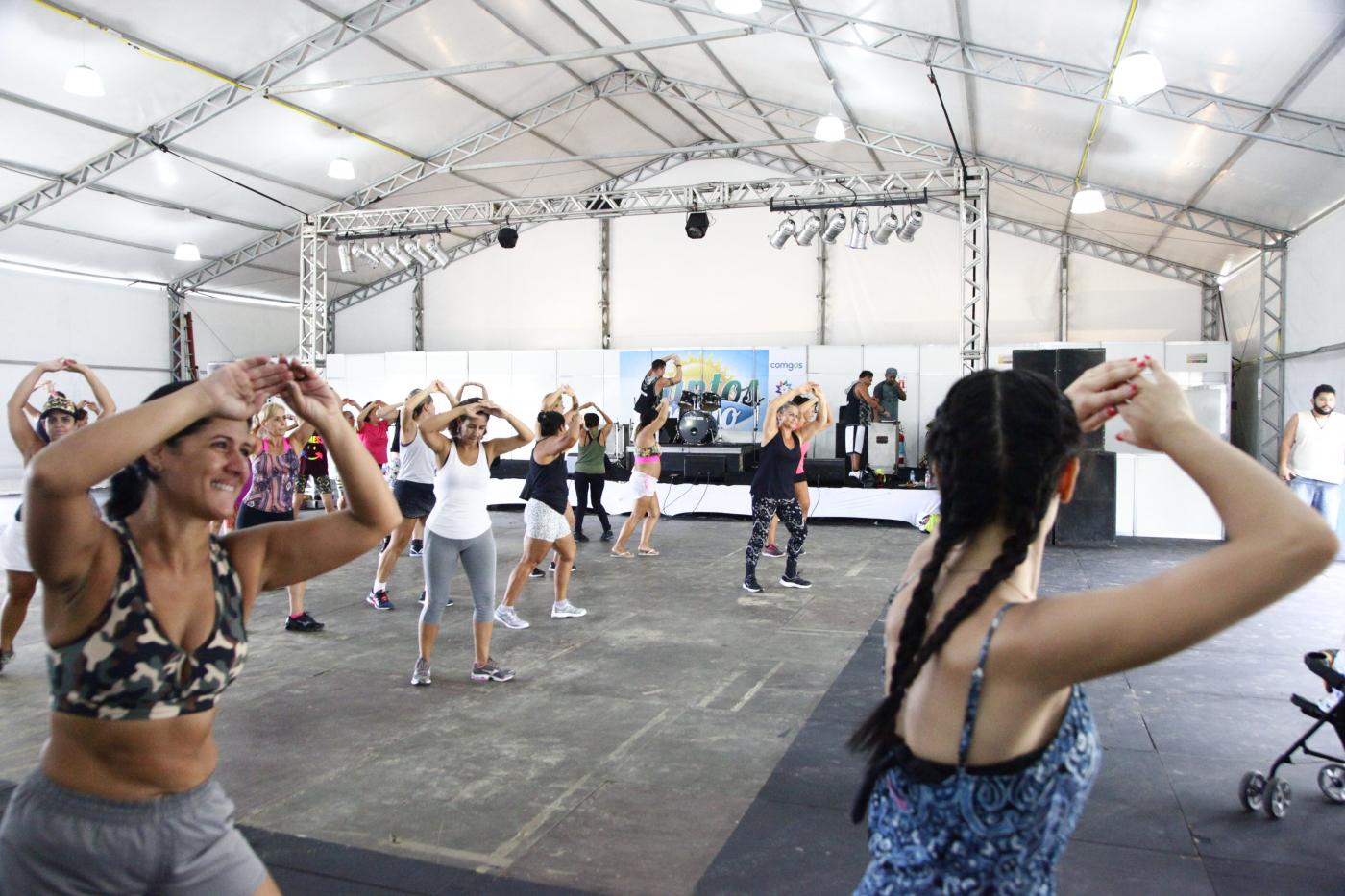 Pessoas dançam sob tenda. Em primeiro plano, duas mulheres estão com braços para o alto. Elas se movimentam. Uma delas sorri. A outra está de costas. #Pracegover