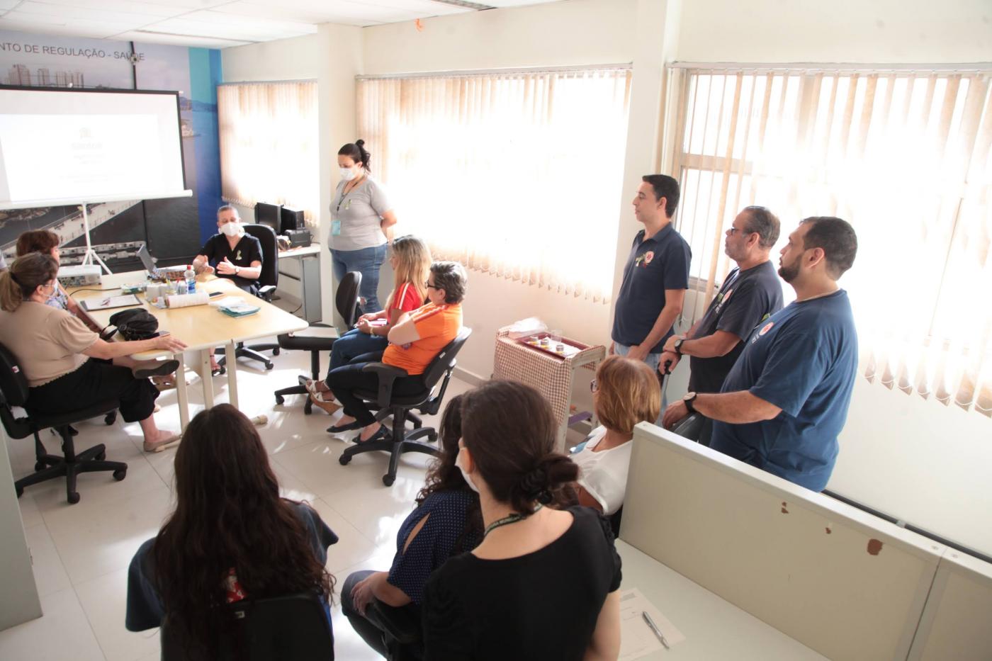 pessoas sentadas em uma sala , mesa ao fundo, à direita três pessoas em pé #paratodosverem