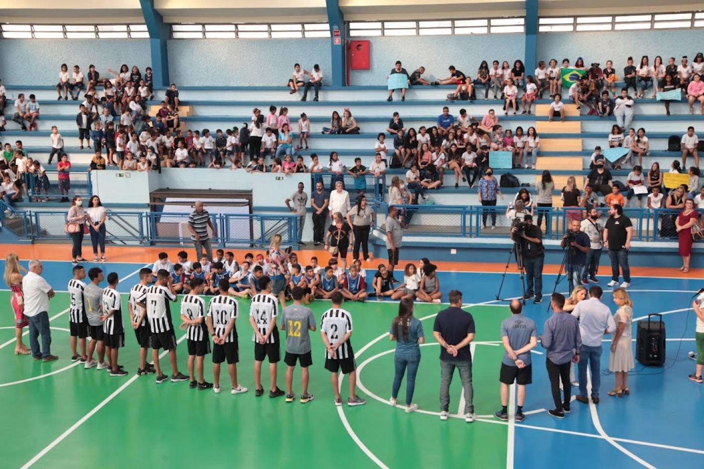 jogadores na quadra e diversos estudantes os olhando na arquibancada #paratodosverem