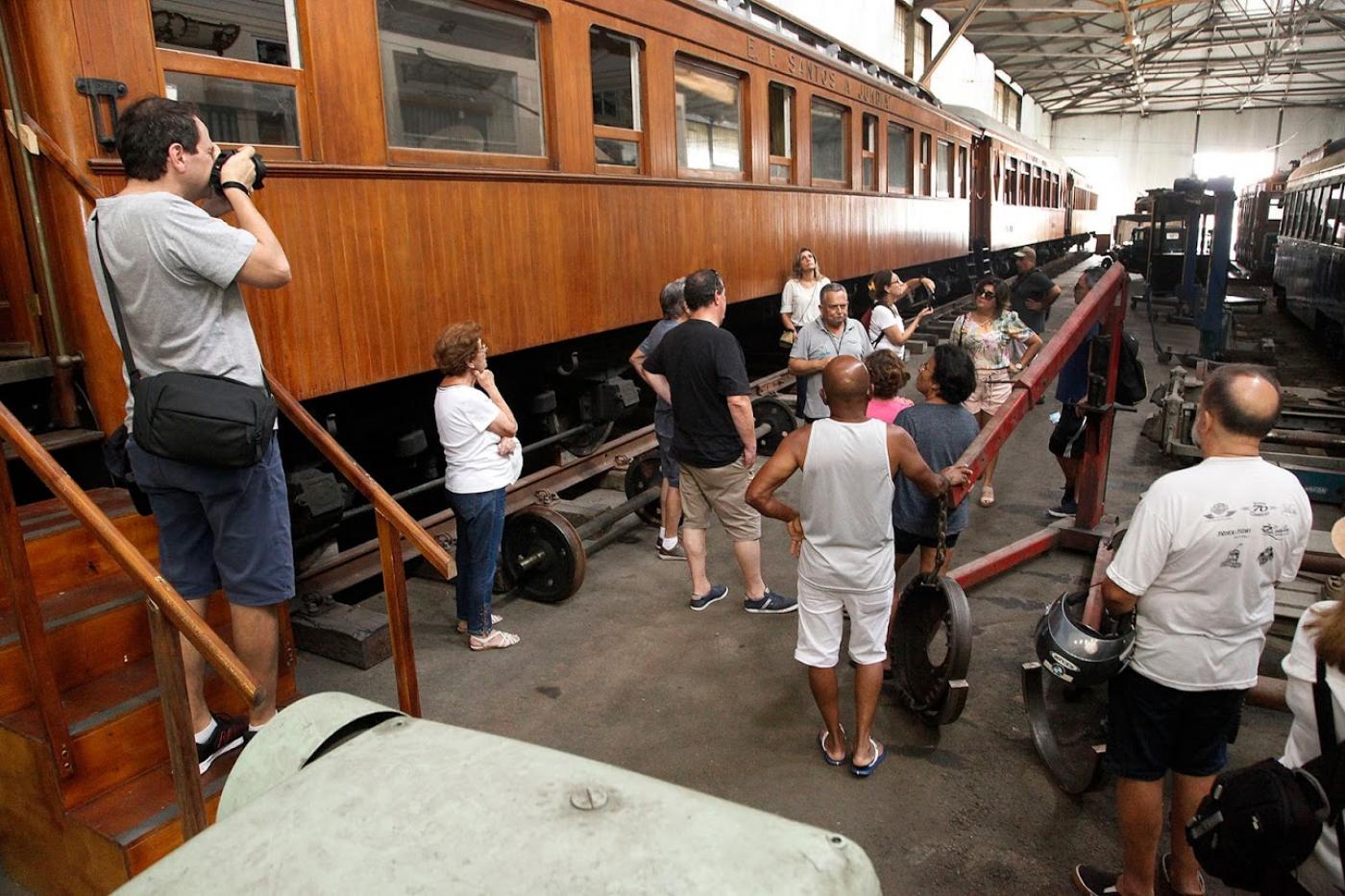 Visitantes na garagem de bonde ouvem explicações de guia de turismo e observam vagões antigos. #Paratodosverem