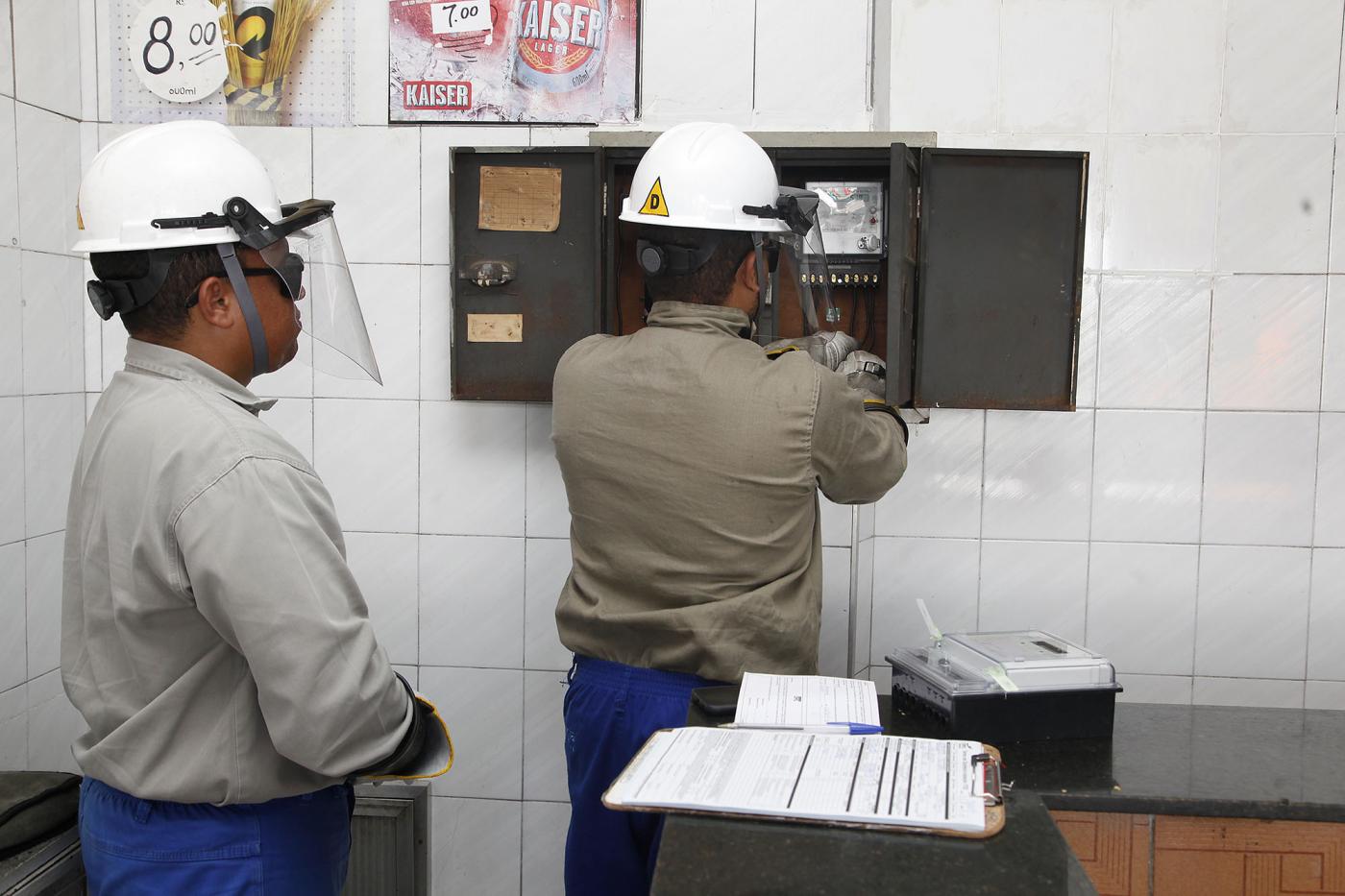 Funcionários da CPFL, com capacete e protetor visual verificam relógio de luz em estabelecimento. #Paratodosverem