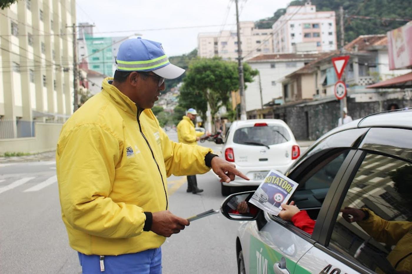 agente da cet uniformizado entrega folheto para pessoa dirigindo um taxo. Outro agente está à frente fazendo o mesmo. #paratodosverem