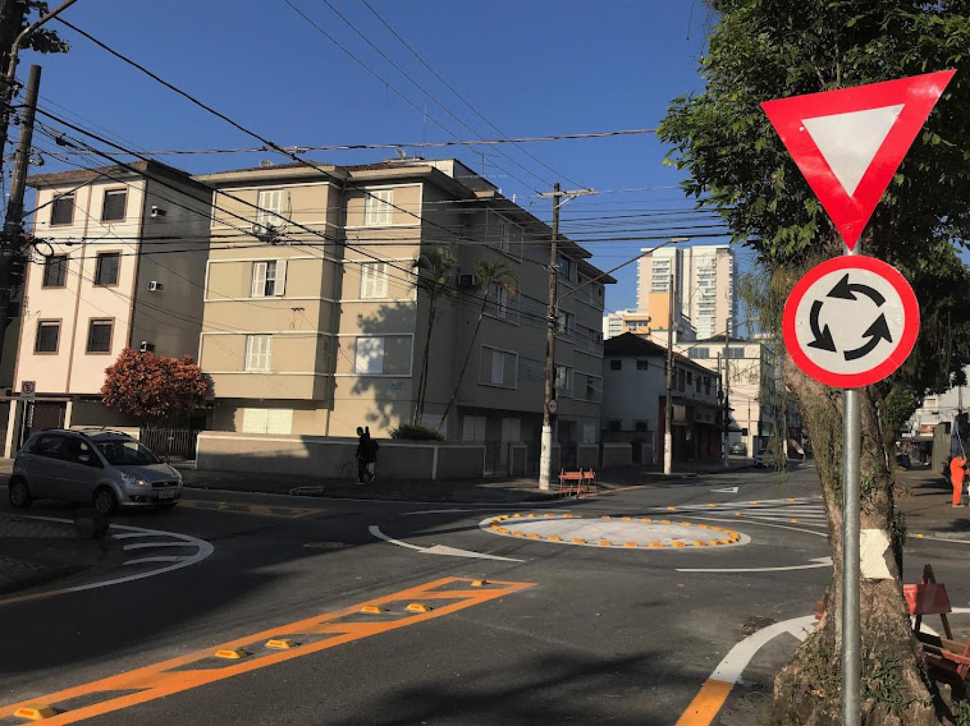 Placa de minirrotatória em primeiro plano, com dispositivo ao fundo durante passagem de veículo. #pracegover